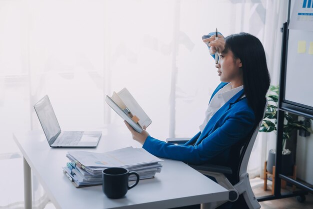 Foto mujer asiática con migraña y tensión en la cabeza mujer de negocios y financista sobrecargada de trabajo mientras trabaja en una computadora portátil y tableta en una oficina moderna