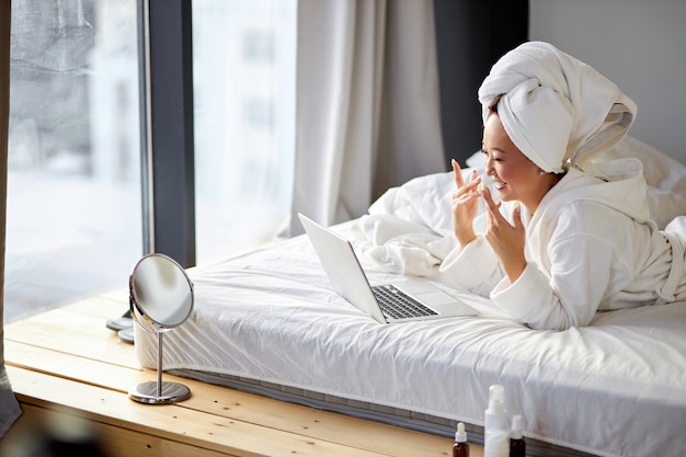 Mujer asiática mira la pantalla de la computadora portátil hablando, sonriendo, tener una conversación en línea con alguien, en casa en la cama