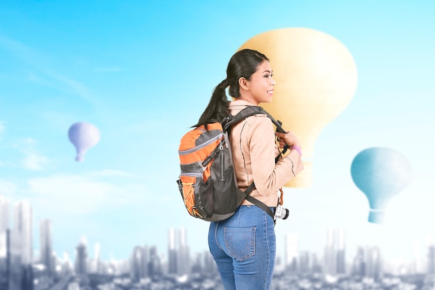 Mujer asiática con mochila mirando coloridos globos volando con fondo de paisaje urbano