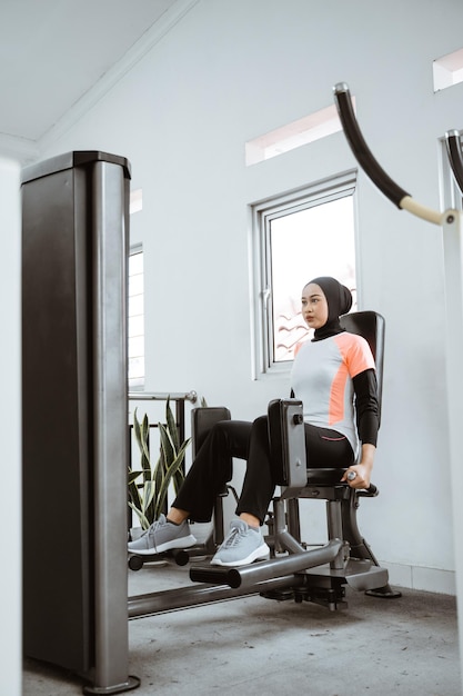 Mujer asiática musulmana trabajando en la parte inferior de su cuerpo usando una máquina de piernas en el gimnasio