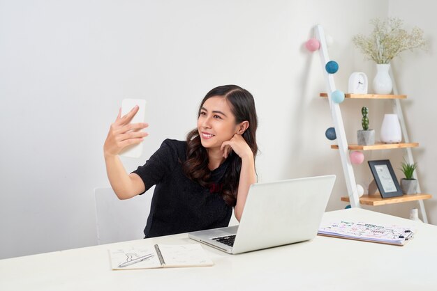 Mujer asiática de negocios casual tomando un selfie con smartphone frente a una computadora portátil en la oficina.