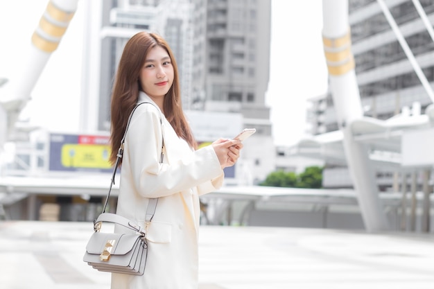 La mujer asiática de negocios joven en un traje crema está usando el teléfono móvil (smartphone) al aire libre para contactar con socios mientras va a trabajar en la oficina.