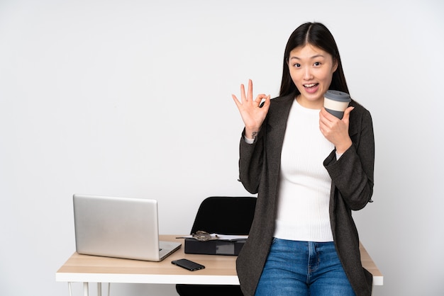 Mujer asiática de negocios en su lugar de trabajo aislado en la pared blanca que muestra signo ok y pulgar arriba gesto