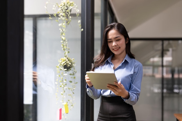 Mujer asiática de negocios usando tableta digital de pie cerca de la ventana en la oficina