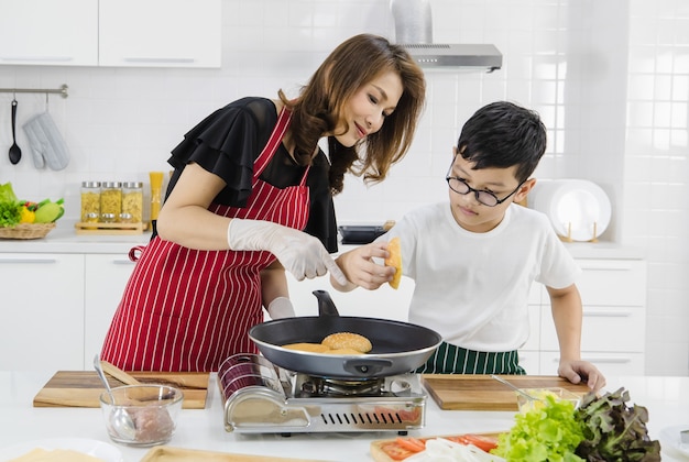 Mujer asiática y niño poniendo bollos frescos en una sartén caliente mientras se cocinan hamburguesas para el almuerzo en la cocina de casa. Conept para compartir el tiempo de amor y relación en la familia moderna.