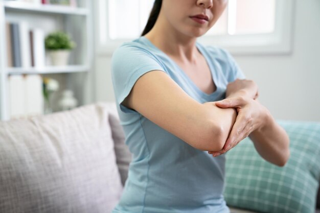 una mujer asiática no reconocida mira el dolor de su codo magullado. ama de casa china casual sosteniendo su codo sentada en el sofá en casa en el interior de una casa moderna y luminosa cerca del sol a través de la ventana.