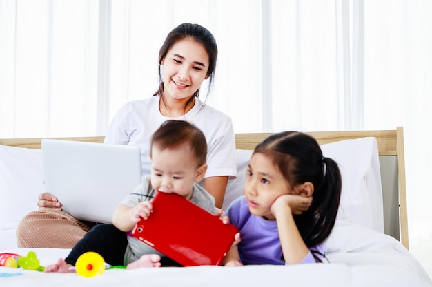 Mujer asiática ocupada cuidando de su bebé y su pequeña hija y trabajando desde casa con una laptop Super mamá