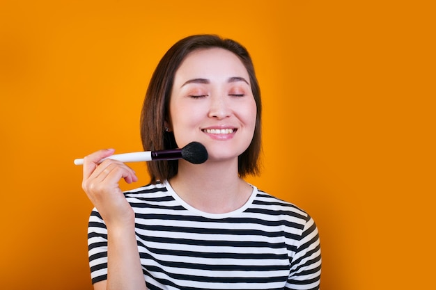 Mujer asiática de pelo corto aplicando cepillo de polvo cosmético sobre fondo amarillo