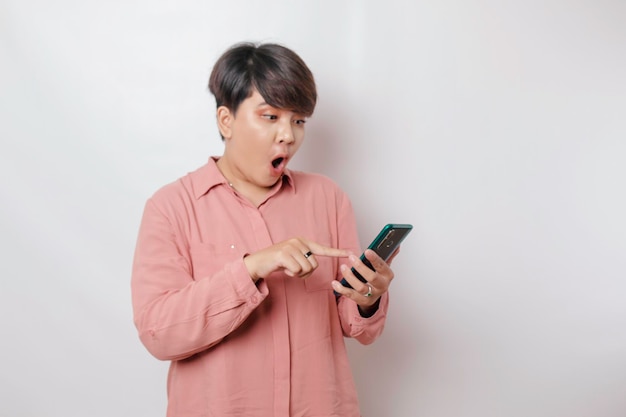 Mujer asiática de pelo corto sorprendida con una camisa rosa apuntando a su teléfono inteligente aislado por un fondo blanco