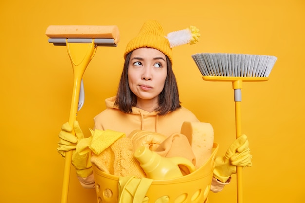 Mujer asiática pensativa cansada lava la ropa en casa tiene herramientas de limpieza ocupadas con la limpieza que va a barrer el baño de lavado de piso usa sudadera de sombrero y guantes de goma aislados sobre fondo amarillo