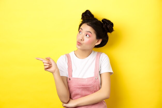 Mujer asiática pensativa con maquillaje de belleza, apuntando y mirando a la izquierda con cara curiosa, interesada en publicidad, de pie en amarillo.