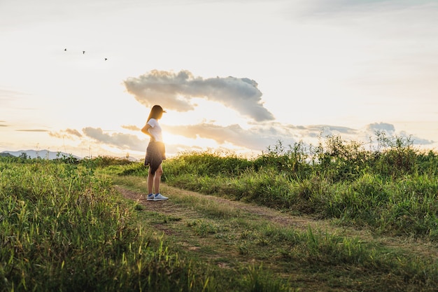 Mujer asiática de pie en el campo de hierba con fondo de paisaje escénico de cielo de montaña y puesta de sol con espacio de copia.