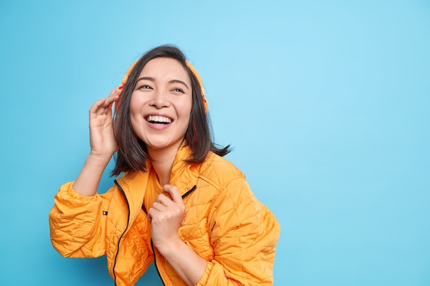 La mujer asiática positiva disfruta de su música favorita en los auriculares, las sonrisas expresan ampliamente la felicidad y usan una chaqueta naranja aislada sobre fondo azul con espacio para copiar su anuncio. Estilo de vida