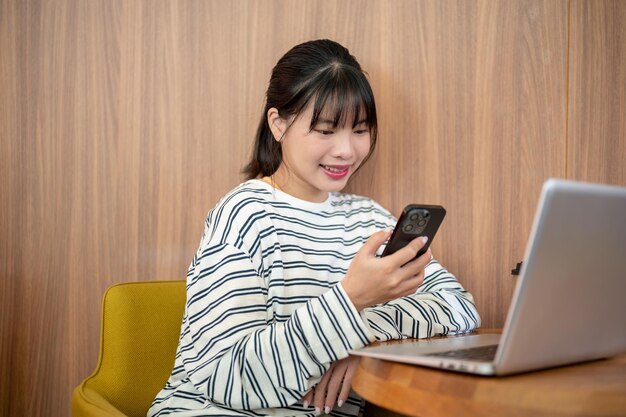 Una mujer asiática positiva está usando su teléfono inteligente mientras está sentada en una mesa con su computadora portátil en la habitación