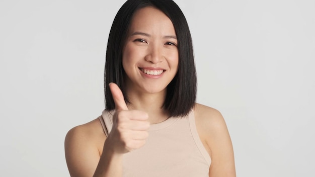 Mujer asiática positiva manteniendo el pulgar hacia arriba sobre fondo gris Chica asiática sonriendo a la cámara mostrando un gesto similar