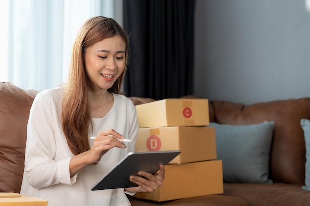 Mujer asiática con propietaria de una pequeña empresa de valores con tableta digital y cajas de paquetes de paquetes minoristas que verifican el pedido de entrega de envío comercial en un teléfono inteligente usando tecnología de aplicación móvil