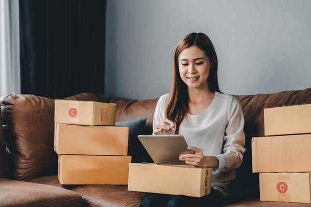 Mujer asiática con propietaria de una pequeña empresa de valores con tableta digital y cajas de paquetes de paquetes minoristas que verifican el pedido de entrega de envío comercial en un teléfono inteligente usando tecnología de aplicación móvil