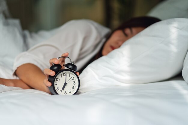 Una mujer asiática que apaga un despertador mientras duerme en una acogedora cama blanca por la mañana
