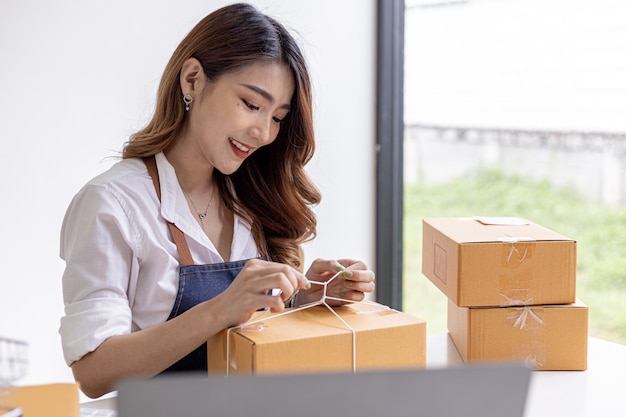 Una mujer asiática que ata un paquete a la caja de un cliente, es dueña de una tienda en línea, empaca y envía a través de una empresa de transporte privada. Conceptos de venta online y compra online.