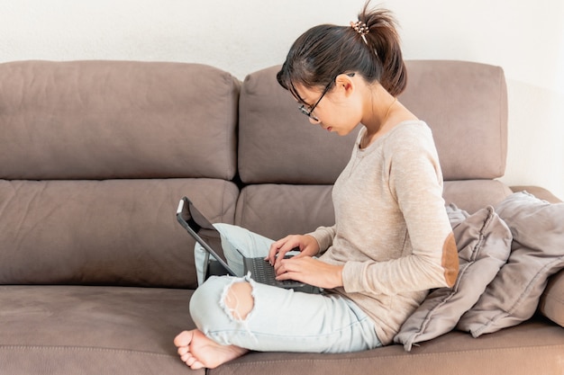 Mujer asiática que trabaja desde casa con una computadora portátil o tableta en el sofá