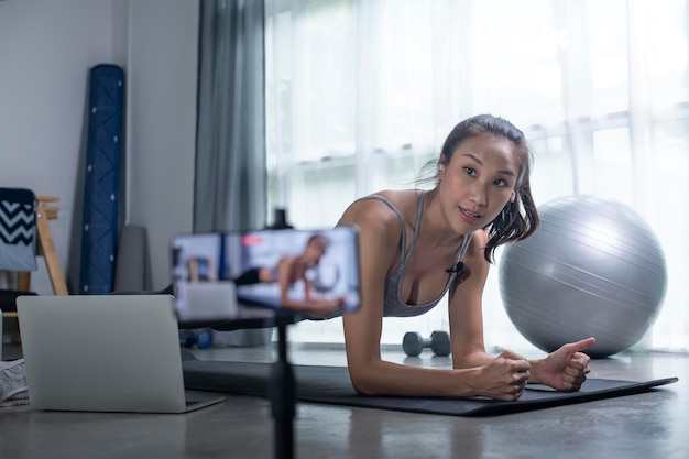 Mujer asiática que usa la cámara del teléfono para transmitir en vivo enseña yoga en línea en la sala de estar mujer joven en ropa deportiva haciendo ejercicio en casa viendo el aula de fitness en línea