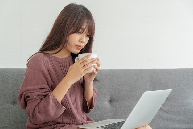Mujer asiática que usa la computadora portátil en el sofá en la sala de estar