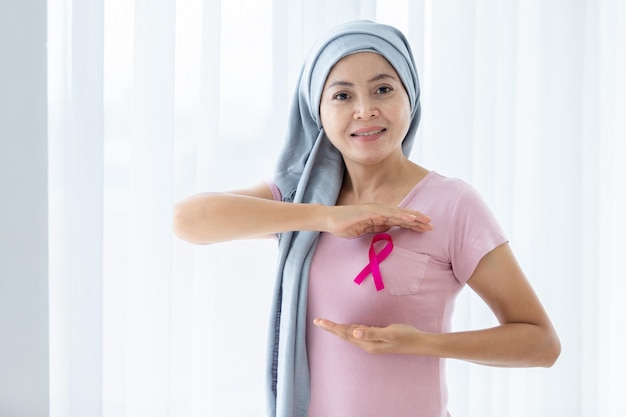Foto una mujer asiática que usa la enfermedad del pañuelo en la cabeza con la mano de cinta rosa revisando bultos en su seno en busca de signos de cáncer de mama aislado en la ventana en el dormitorio de la medicina de atención médica de la casa