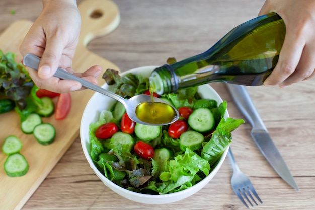 Mujer asiática que vierte el aceite de oliva en el tazón de ensalada de verduras frescas en la cocina