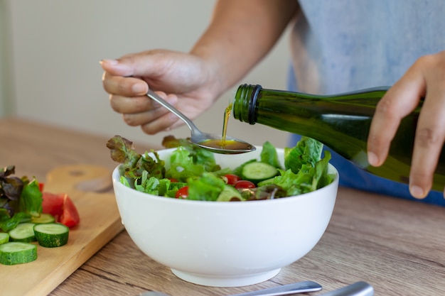 Foto mujer asiática que vierte el aceite de oliva en el tazón de ensalada de verduras frescas en la cocina