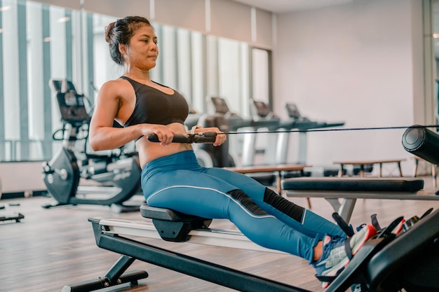 Una mujer asiática quema calorías en una máquina de remo en un gimnasio deportivo. Ella es deliberadamente para abdominales fuertes. Lleva una barra deportiva negra y pantalones azules. FitnessGymsport y concepto de estilo de vida saludable