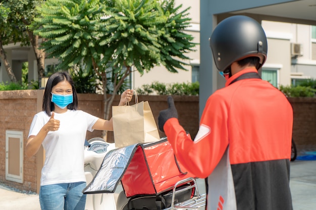 La mujer asiática recoge la bolsa de comida de entrega de la caja y el pulgar forma sin contacto o sin contacto del jinete de entrega con la bicicleta en la casa para el distanciamiento social por riesgo de infección. Concepto de coronavirus