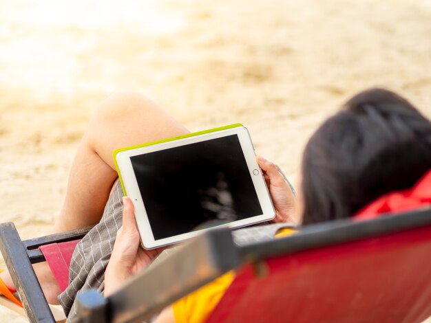 La mujer asiática se relaja en la cama de la playa y juega con la tableta