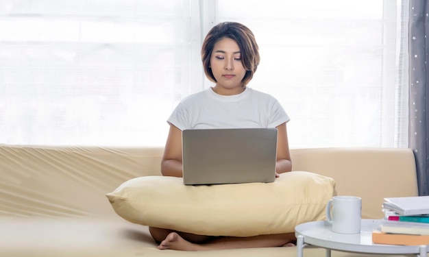 La mujer asiática se relaja trabajando con una computadora portátil en el sofá en casa como trabajo en casa en una pandemia