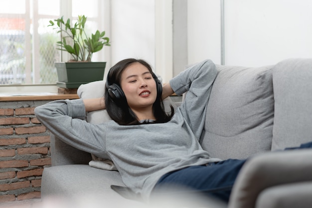 Mujer asiática relajada en música que escucha de los auriculares y que coloca en el sofá.