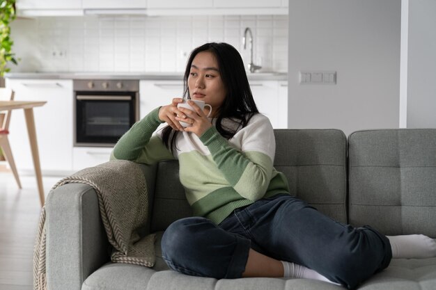 Una mujer asiática relajada y pensativa con una taza de té y café soñadora mirando hacia otro lado se sienta en el sofá en la sala de estar