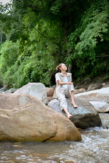 Mujer asiática relajada sentada cerca del río en la piedra del río tomando aire fresco acampando