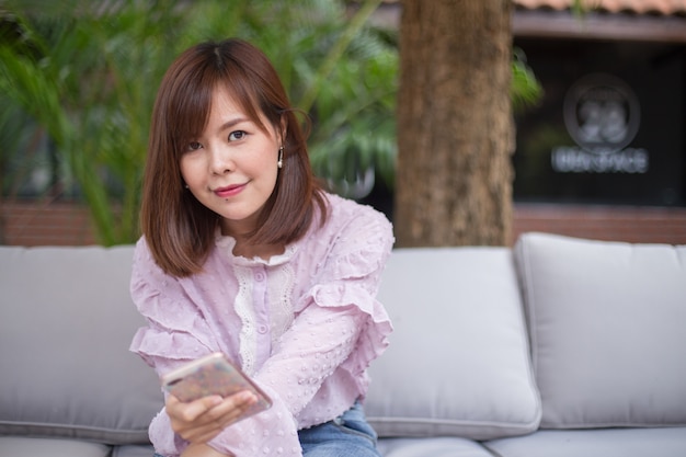 Mujer asiática del retrato que usa un teléfono elegante en jardín de la cafetería