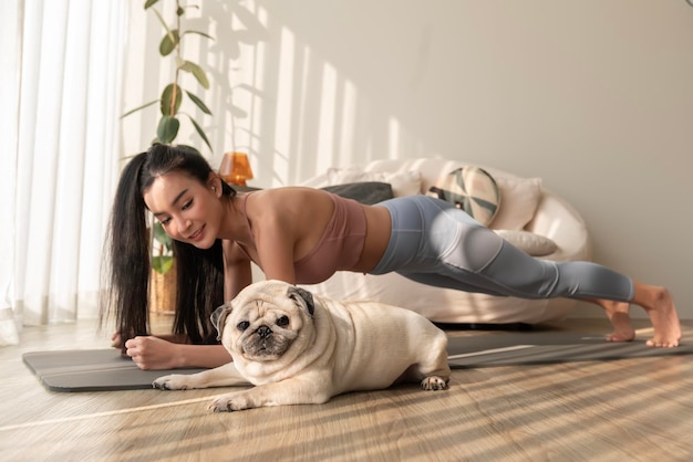 Mujer asiática en ropa deportiva haciendo ejercicio y haciendo yoga con un lindo perro en la sala de estar de casa