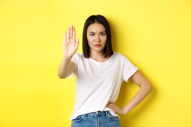 La mujer asiática segura y seria dice que no, mostrando un gesto de parada para prohibir y advertir, en desacuerdo con alguien, de pie molesto contra el fondo amarillo.
