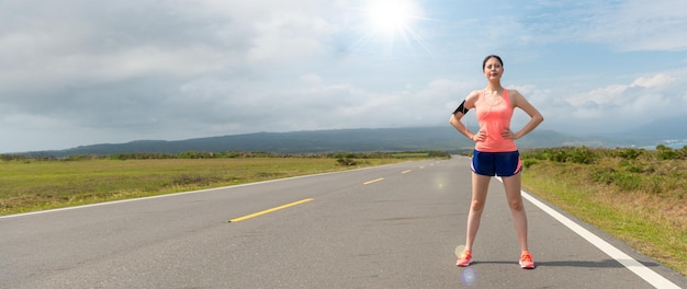 mujer asiática segura de sí misma parada en la carretera lista para correr para entrenar resistencia en la tarde del día soleado con un hermoso paisaje de fondo. Recorte de banner para espacio de copia.