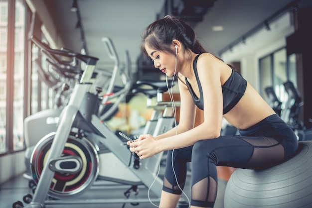 Mujer asiática sentada en la bola de la aptitud y escuchar música en el gimnasio de entrenamiento de fitness