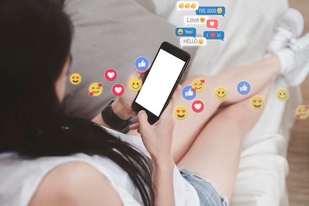 Foto mujer asiática sentada en un sofá relajándose con la pantalla blanca del teléfono celular mujer joven relajándose con las redes sociales solo mujeres usando el teléfono inteligente sosteniendo y escribiendo con otros a través de emoji y texto en línea