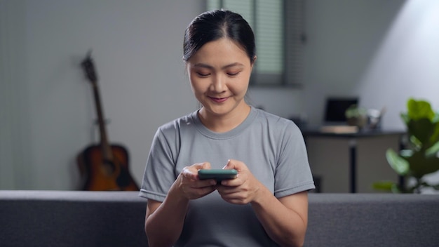 Mujer asiática sentada en un sofá y sosteniendo un teléfono inteligente en casa Mujer feliz usando un teléfono inteligente para buscar compras en línea o navegar en las redes sociales