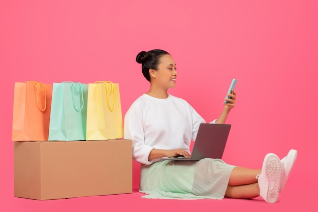 Mujer asiática sentada en el suelo entre bolsas de compras usando una computadora portátil y un teléfono inteligente