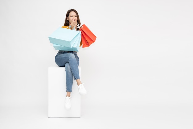 mujer asiática, sentado, en, caja blanca, y, tenencia, bolsas de compras, aislado, blanco, plano de fondo