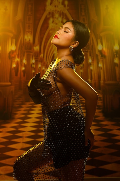 Mujer asiática sexy de los años 20 con hermosa cara de pecas y cuerpo regordete con un vestido de brillo sensual de cristal sobre luz amarilla dorada en la mansión Palace para iluminación de estudio de fiesta privada