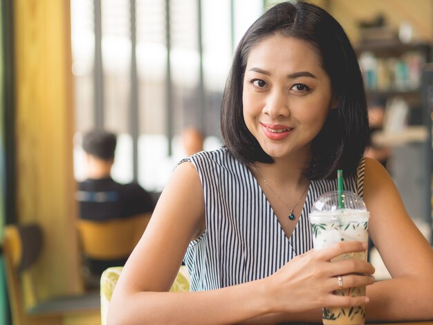 La mujer asiática se sienta en café del café wirh helado sobre la mesa.