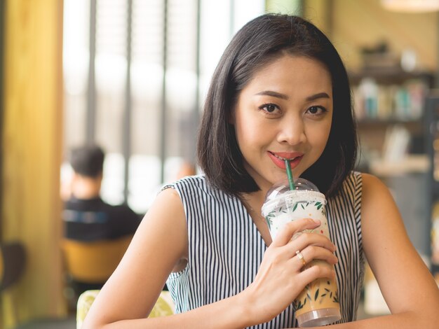 La mujer asiática se sienta en café del café wirh helado sobre la mesa.