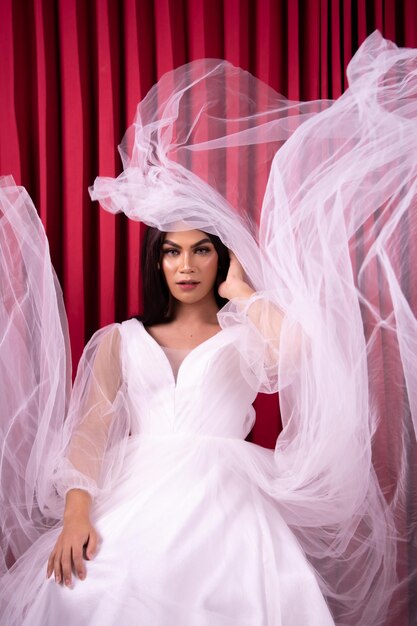 La mujer asiática se sienta con glamour mientras el vestido de novia vuela a su alrededor frente a la cortina roja
