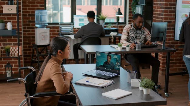 Mujer asiática en silla de ruedas reunida con un hombre en una videoconferencia, planificando el crecimiento empresarial en una teleconferencia en línea. Trabajador de oficina que usa videoconferencia remota en una computadora portátil.